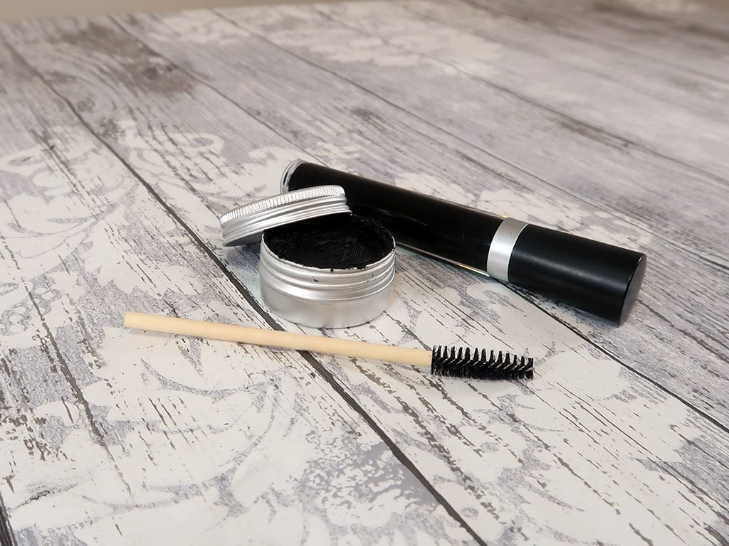 Two beautiful mascaras, one in a tube and the other in a pot, next to an applicator brush